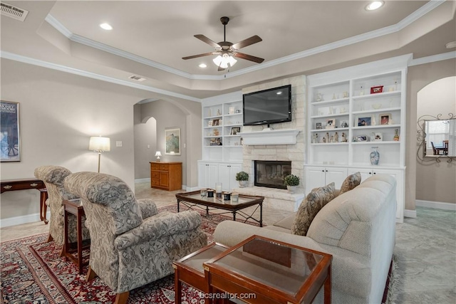 living room with a raised ceiling, a stone fireplace, ceiling fan, and crown molding