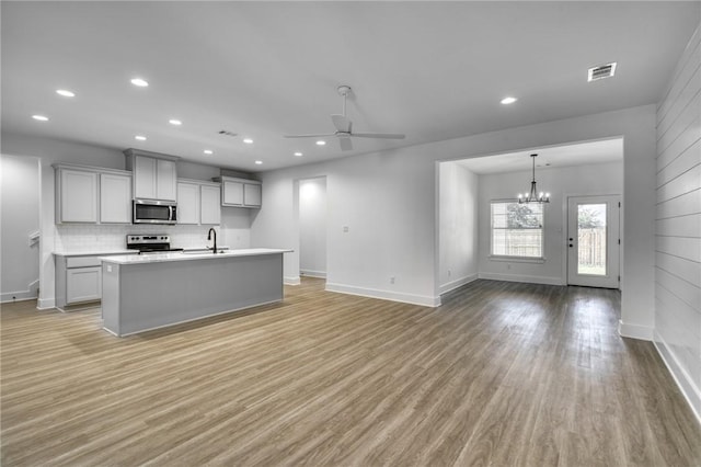 kitchen with ceiling fan with notable chandelier, stainless steel appliances, sink, light hardwood / wood-style flooring, and an island with sink