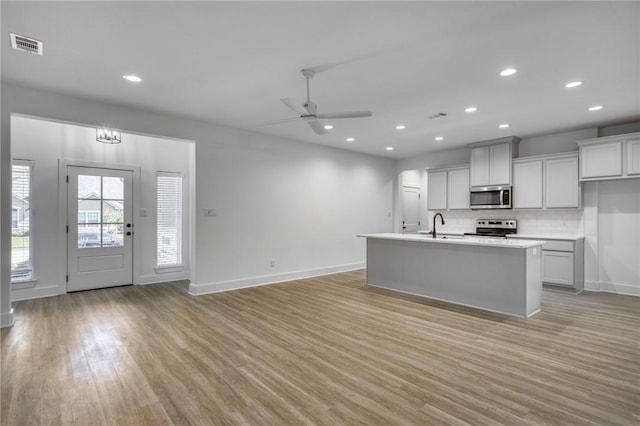 kitchen with ceiling fan, stainless steel appliances, tasteful backsplash, an island with sink, and light wood-type flooring