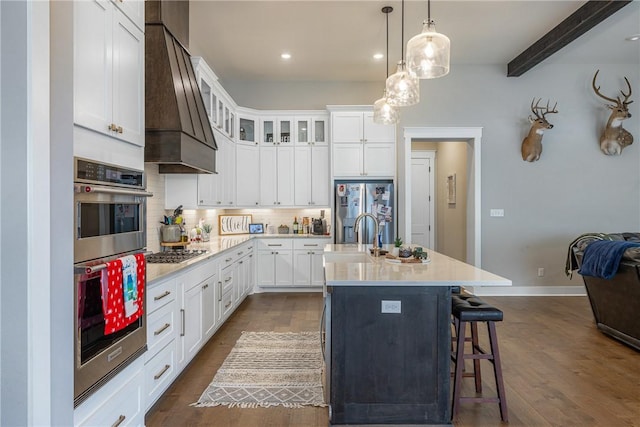 kitchen featuring beam ceiling, stainless steel appliances, dark hardwood / wood-style flooring, premium range hood, and an island with sink