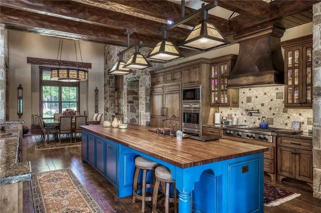 kitchen featuring premium range hood, built in appliances, beamed ceiling, a kitchen island, and butcher block counters