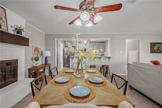 tiled dining space featuring a brick fireplace, ceiling fan, and ornamental molding