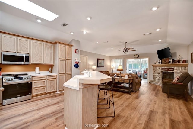 kitchen featuring a center island with sink, a fireplace, light brown cabinetry, light hardwood / wood-style floors, and stainless steel appliances