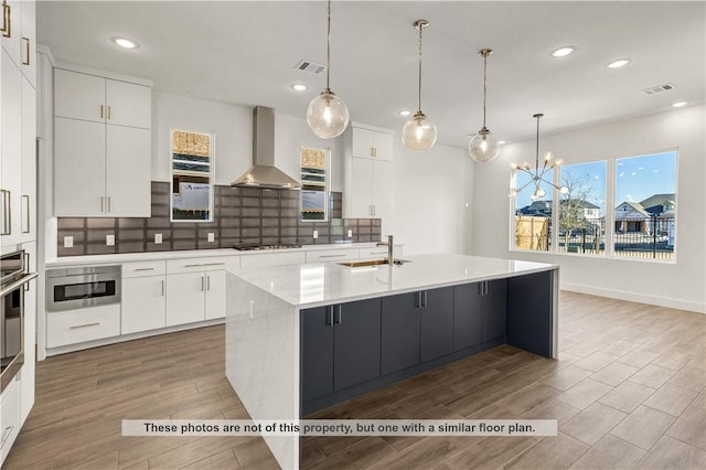 kitchen with wall chimney exhaust hood, hanging light fixtures, a large island with sink, light hardwood / wood-style floors, and appliances with stainless steel finishes