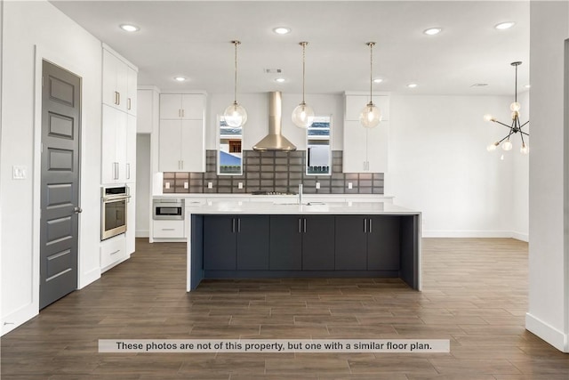 kitchen featuring appliances with stainless steel finishes, wall chimney exhaust hood, a large island with sink, white cabinets, and hanging light fixtures