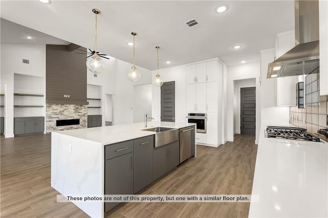 kitchen with high vaulted ceiling, white cabinets, a center island with sink, wall chimney range hood, and appliances with stainless steel finishes