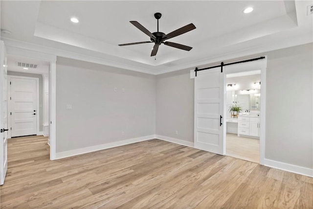 empty room with a barn door, light hardwood / wood-style flooring, ceiling fan, and a tray ceiling