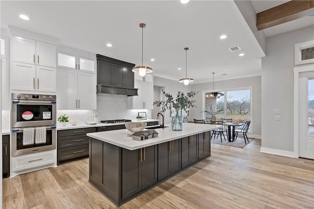 kitchen with appliances with stainless steel finishes, a kitchen island with sink, white cabinetry, light hardwood / wood-style floors, and decorative light fixtures