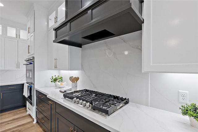 kitchen featuring wall chimney exhaust hood, light stone counters, white cabinets, stainless steel appliances, and backsplash