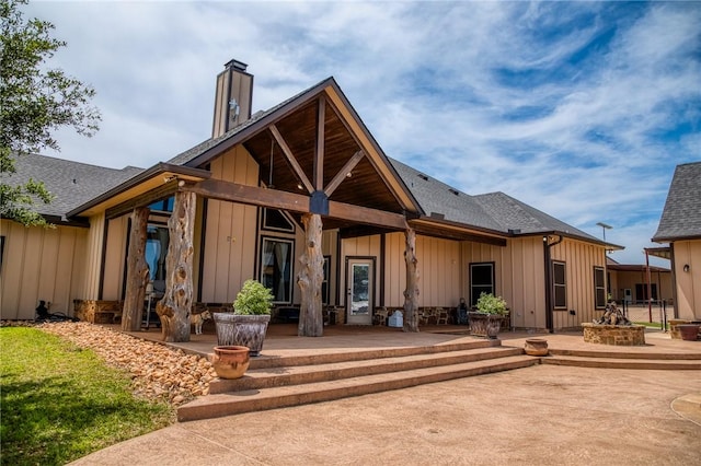 rear view of property with board and batten siding, a chimney, an outdoor fire pit, a patio, and a shingled roof
