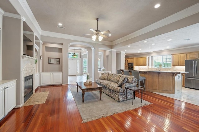 living area with visible vents, ceiling fan, light wood-type flooring, a tile fireplace, and arched walkways