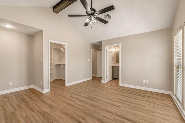 unfurnished bedroom featuring a walk in closet, light wood-type flooring, ceiling fan, ensuite bathroom, and beam ceiling