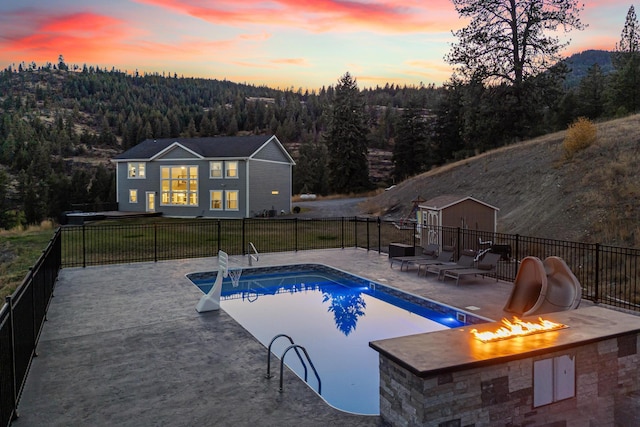 pool at dusk with a fire pit, a lawn, a water slide, a patio area, and an outbuilding