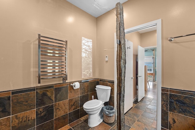 bathroom featuring toilet, a wainscoted wall, tile walls, radiator, and stone tile flooring