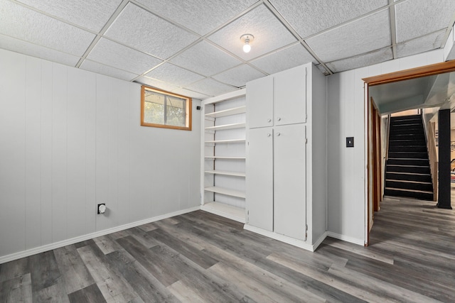 below grade area with a paneled ceiling, baseboards, dark wood finished floors, and stairs