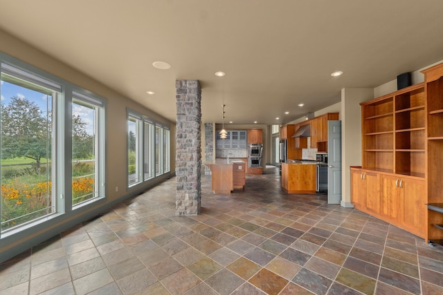 kitchen featuring ornate columns, a kitchen island, double oven, decorative light fixtures, and wall chimney exhaust hood