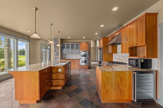 kitchen featuring wall chimney range hood, sink, appliances with stainless steel finishes, beverage cooler, and a large island with sink