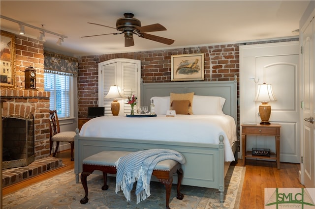 bedroom with ceiling fan, a brick fireplace, light hardwood / wood-style flooring, brick wall, and track lighting