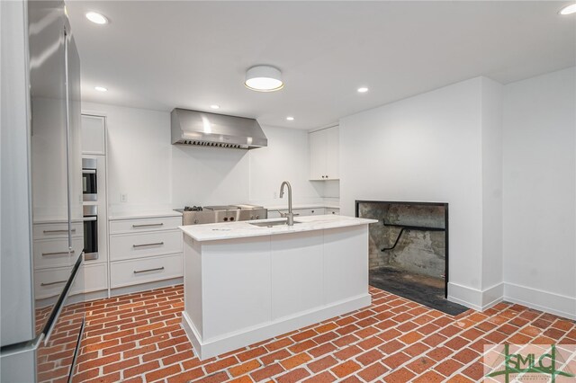 kitchen with white cabinets, wall chimney exhaust hood, sink, and a kitchen island with sink