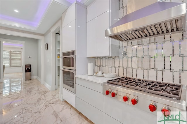 kitchen with wall chimney range hood, light tile flooring, tasteful backsplash, white cabinets, and a tray ceiling