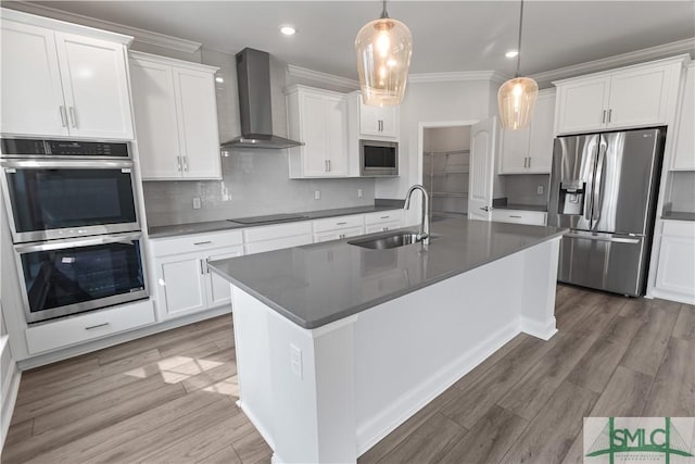 kitchen with a center island with sink, dark countertops, appliances with stainless steel finishes, a sink, and wall chimney range hood