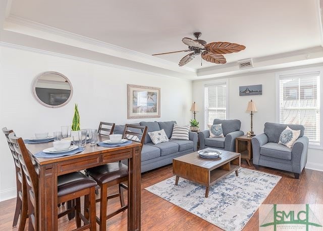 living room with a tray ceiling, wood finished floors, visible vents, ceiling fan, and ornamental molding