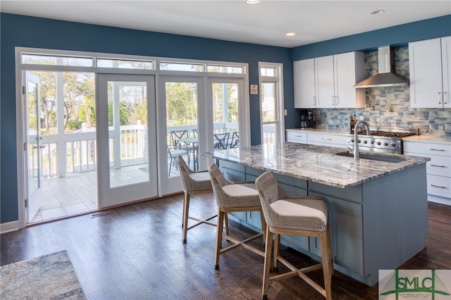 kitchen with wall chimney range hood, dark wood-type flooring, a breakfast bar, white cabinetry, and light stone countertops