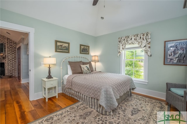 bedroom with ceiling fan, wood-type flooring, vaulted ceiling, and a brick fireplace