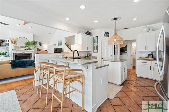 kitchen featuring a brick fireplace, vaulted ceiling, an island with sink, white cabinets, and pendant lighting