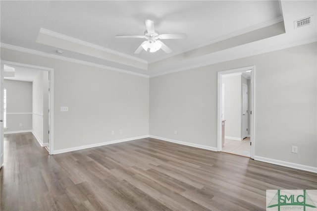 empty room with a ceiling fan, wood finished floors, baseboards, ornamental molding, and a raised ceiling