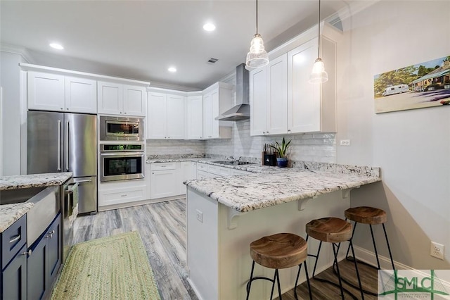 kitchen with appliances with stainless steel finishes, a kitchen breakfast bar, wall chimney exhaust hood, white cabinetry, and hanging light fixtures