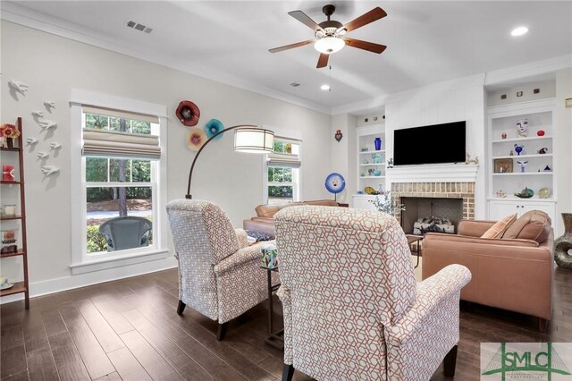 living room featuring built in shelves, ceiling fan, a brick fireplace, dark hardwood / wood-style floors, and crown molding