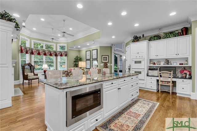 kitchen featuring ceiling fan, light hardwood / wood-style floors, a center island with sink, stainless steel microwave, and white cabinetry