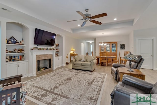 living room featuring a fireplace, built in shelves, ceiling fan with notable chandelier, and a raised ceiling