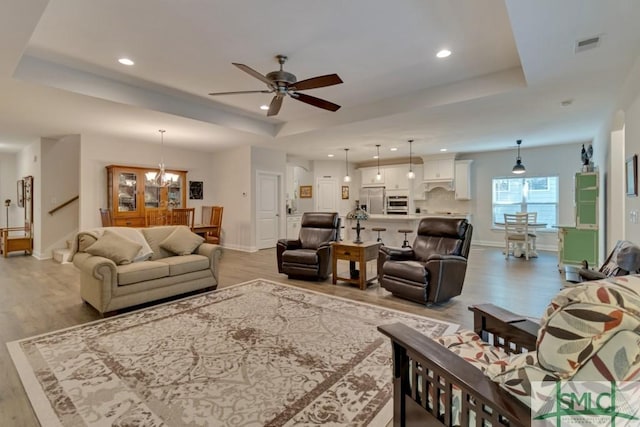 living room featuring ceiling fan with notable chandelier, a tray ceiling, and light hardwood / wood-style flooring