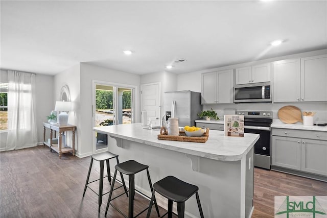 kitchen with gray cabinets, appliances with stainless steel finishes, a breakfast bar, dark hardwood / wood-style flooring, and a kitchen island with sink