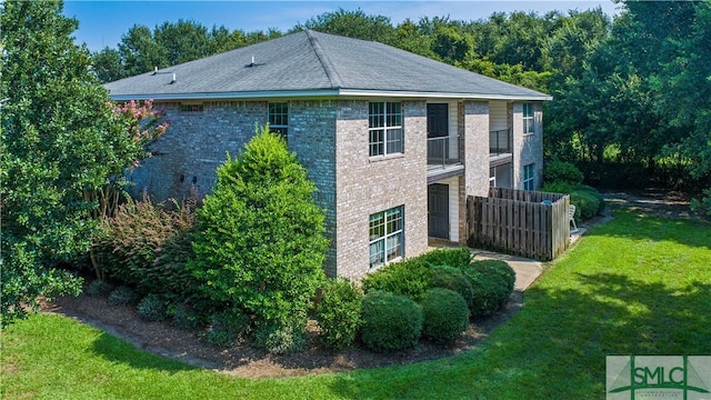 view of home's exterior featuring a balcony, brick siding, fence, roof with shingles, and a lawn