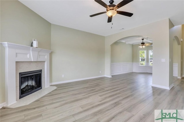 unfurnished living room with arched walkways, ceiling fan, a tile fireplace, wainscoting, and light wood finished floors