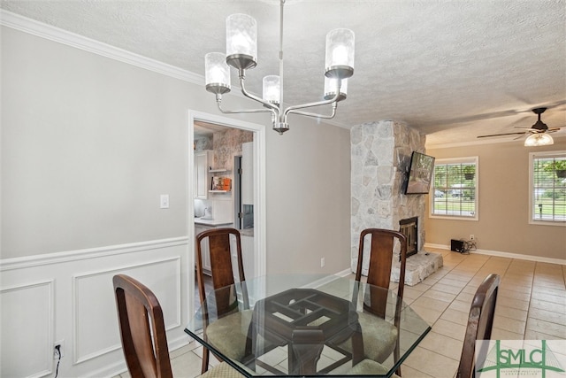 tiled dining room featuring ceiling fan with notable chandelier, a textured ceiling, ornamental molding, and a stone fireplace