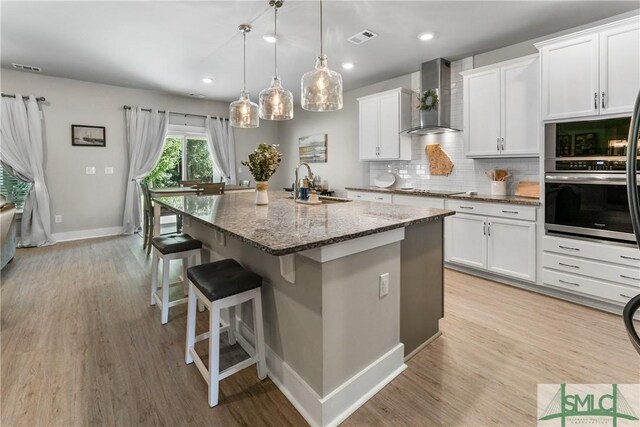 kitchen featuring light wood-type flooring, a center island with sink, stainless steel oven, and wall chimney range hood