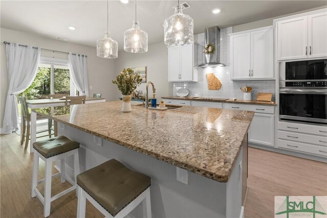 kitchen featuring tasteful backsplash, black appliances, sink, a center island with sink, and wall chimney range hood