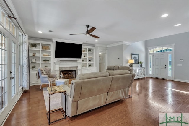 living room with a tile fireplace, built in features, crown molding, dark wood-type flooring, and ceiling fan