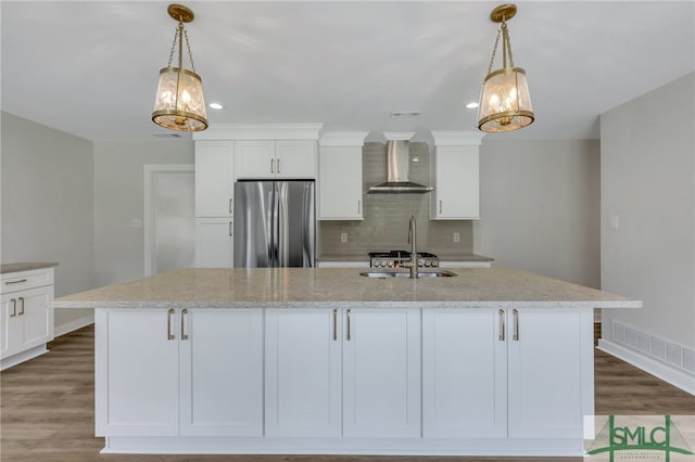 kitchen featuring an island with sink, stainless steel refrigerator, wall chimney range hood, and tasteful backsplash