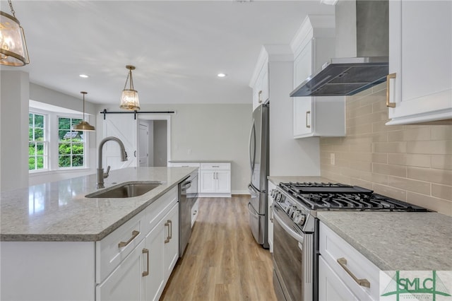 kitchen with light wood-type flooring, stainless steel appliances, sink, a barn door, and wall chimney range hood