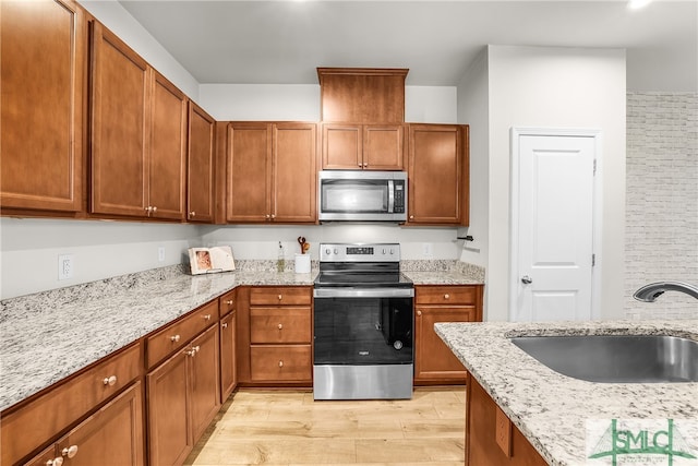 kitchen with light stone counters, brown cabinetry, a sink, light wood-style floors, and appliances with stainless steel finishes