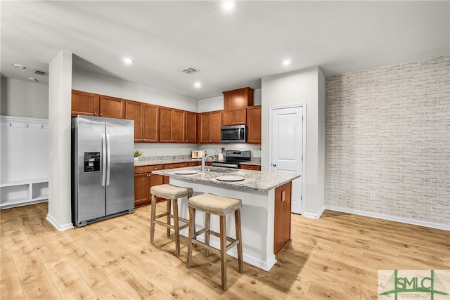 kitchen featuring light stone counters, a center island with sink, brick wall, light wood-style floors, and appliances with stainless steel finishes