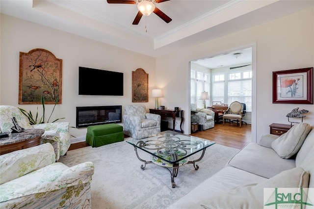 living room with a ceiling fan, a glass covered fireplace, ornamental molding, wood finished floors, and a tray ceiling