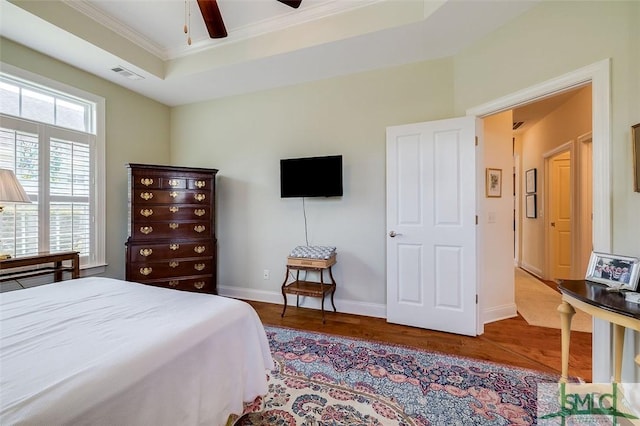 bedroom featuring wood finished floors, visible vents, baseboards, a tray ceiling, and crown molding