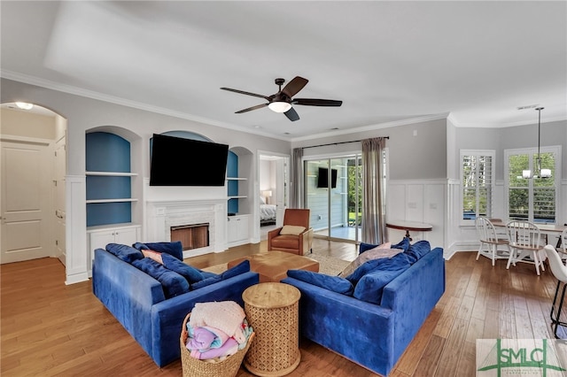 living room with light wood-type flooring, crown molding, ceiling fan with notable chandelier, built in features, and a premium fireplace