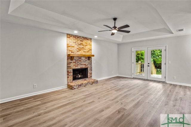 unfurnished living room featuring a brick fireplace, ceiling fan, a tray ceiling, french doors, and light hardwood / wood-style floors